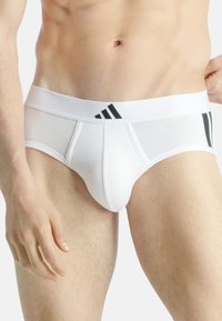 Bañador de algodón blanco con acentos negros, que presenta una cintura elástica ancha con tres rayas negras y un bolsa frontal para soporte.