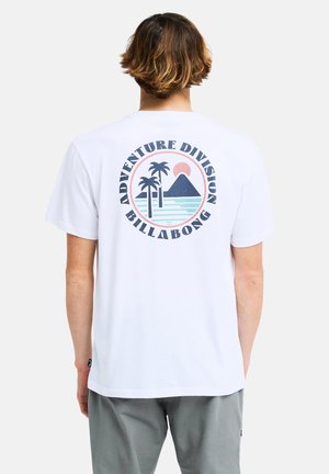 Homme portant un t-shirt blanc avec un logo de plage circulaire montrant deux palmiers, une montagne et le soleil avec le texte Adventure Division Billabong.
