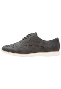 Chaussure en cuir gris avec des détails brogues, à bout rond et semelle en caoutchouc blanche. Elle présente des perforations et six œillets pour les lacets.