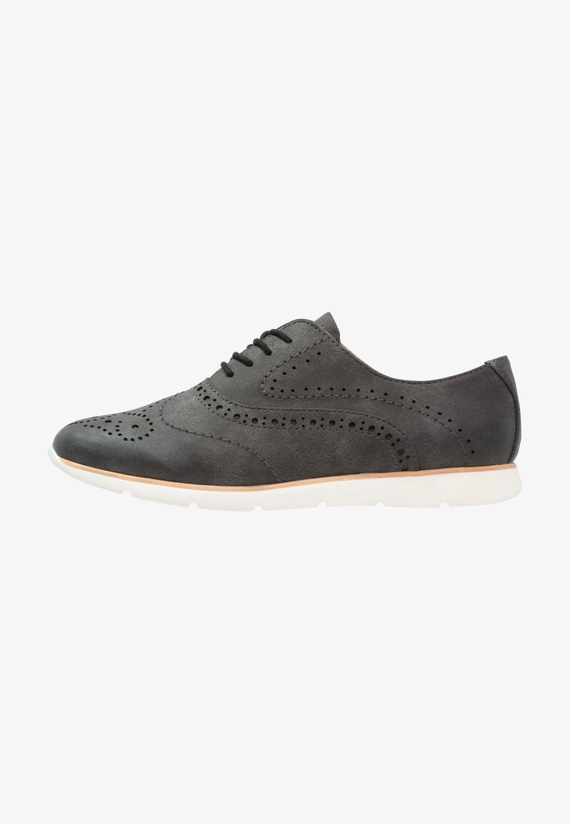Chaussure en cuir gris avec des détails brogues, à bout rond et semelle en caoutchouc blanche. Elle présente des perforations et six œillets pour les lacets.