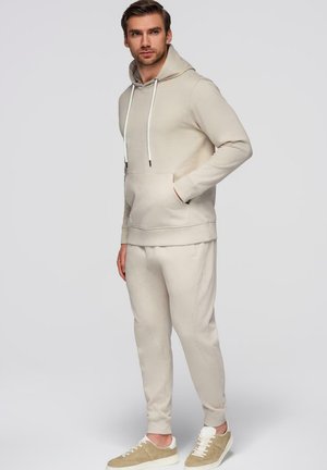 Homme portant un sweat à capuche beige et un pantalon de jogging assorti avec des baskets beige clair, debout devant un fond uni clair.