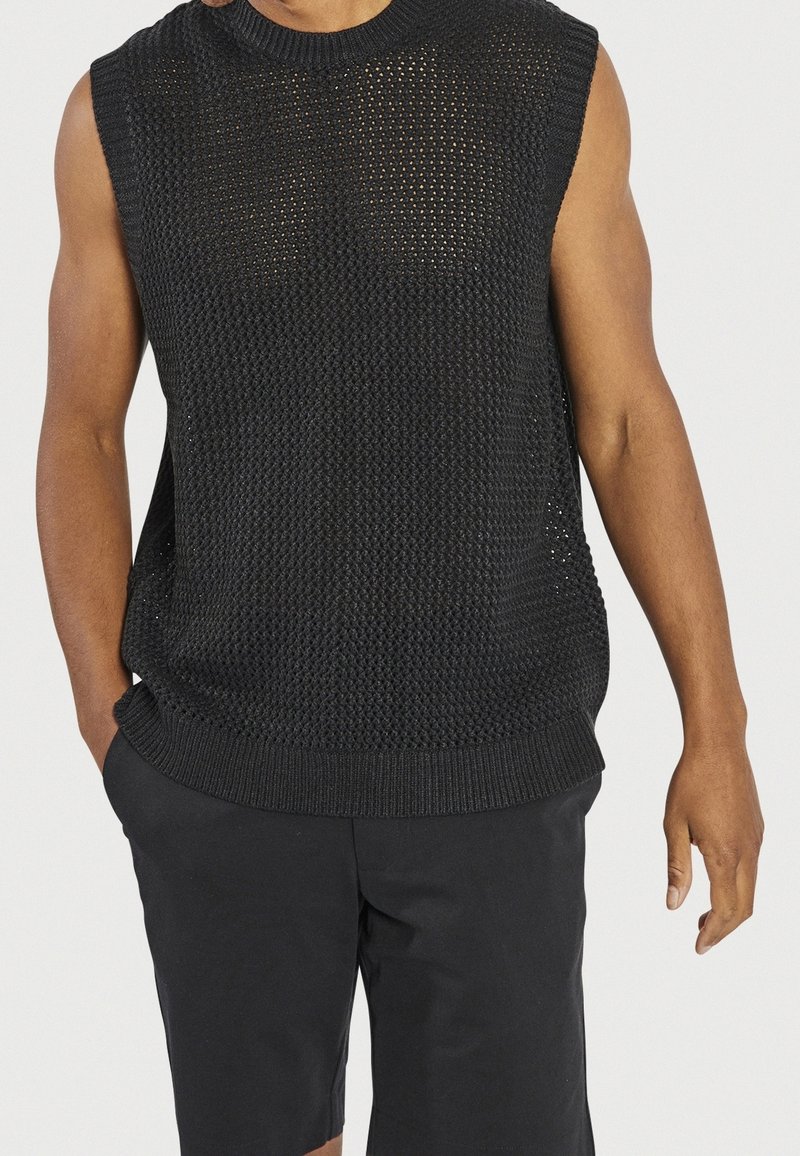 Suéter negro sin mangas de punto con un patrón texturizado, cuello redondo y dobladillo acanalado, combinado con pantalones cortos oscuros.