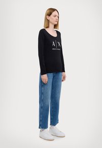 Zwarte longsleeve top met witte "AX" en "Armani Exchange" tekst, gecombineerd met lichtblauwe straight-leg jeans en witte sneakers.