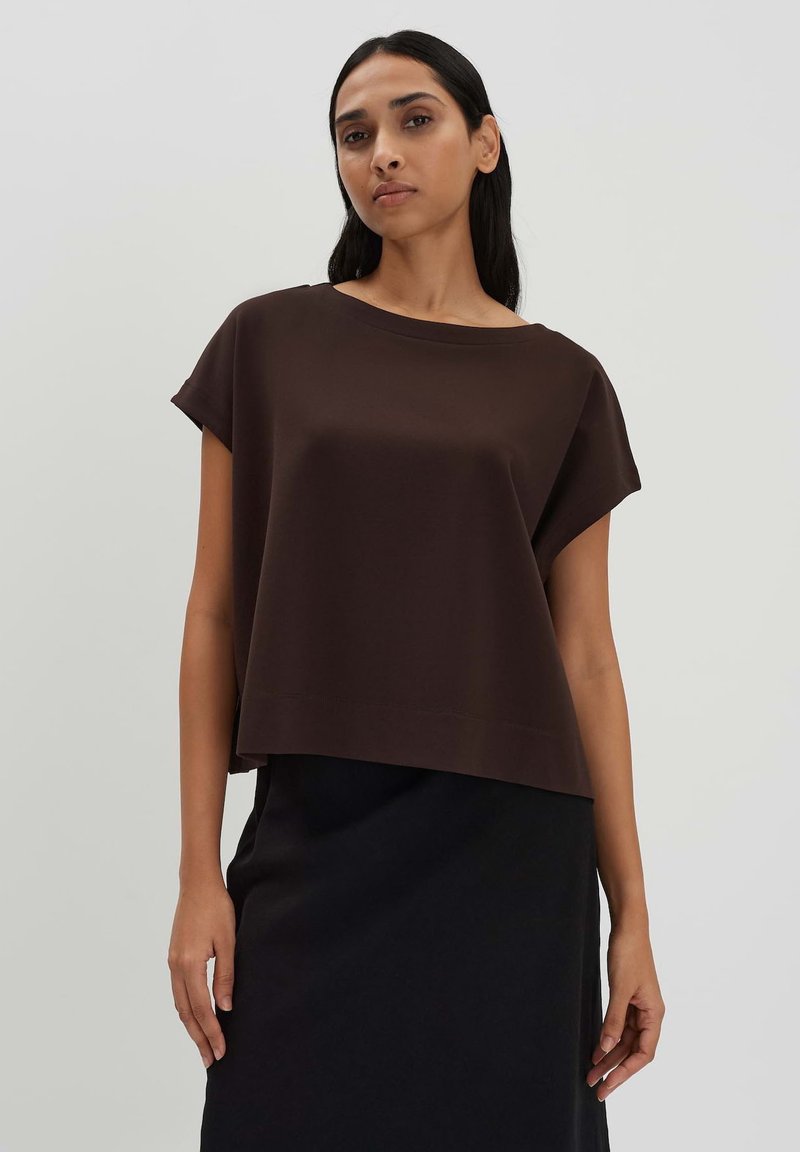 Brauner Cropped-Top mit lockerer Passform, rundem Ausschnitt und kurzen Ärmeln, kombiniert mit einem schwarzen Rock. Glatte Stofftextur.