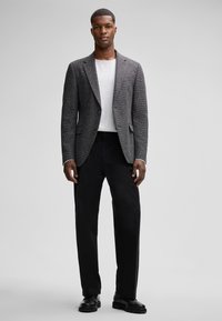 Grau strukturiertes Blazer mit Revers, getragen über einem weißen Hemd und schwarzen Hosen, kombiniert mit schwarzen Lederstiefeln.