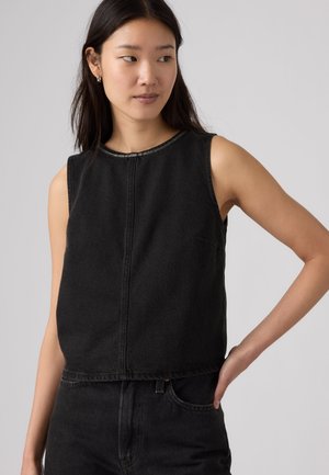 Jonge vrouw met zwarte mouwloze top en zwarte broek kijkt opzij met één hand op haar heup tegen een effen achtergrond.