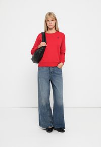 Felpa rossa con collo a giro e piccolo logo a forma di cuore, abbinata a jeans larghi blu. L'individuo tiene una borsa a tracolla nera.
