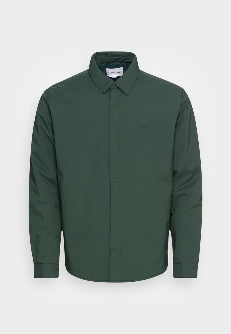 Chemise à manches longues vert foncé avec col et boutons, avec un logo discret sur la poitrine gauche, présentée sur un fond uni.