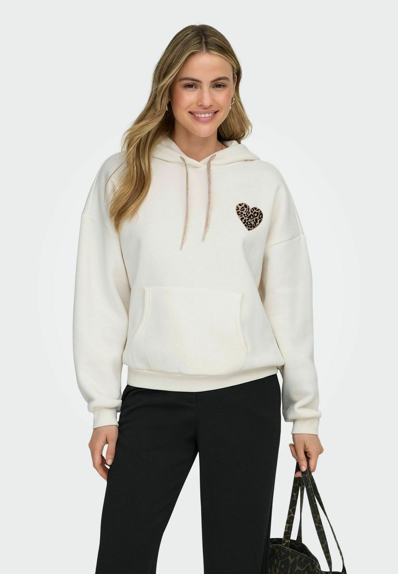 Kremhvit hettegenser med leopardmønster hjerte, snøring og frontlomme. Kombinert med svarte bukser og holder en mønstrete veske.