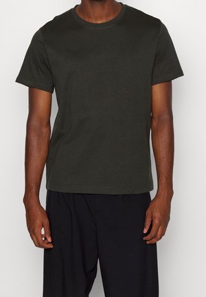 Mand iført en mørkegrøn kortærmet t-shirt med rund hals og løse sorte bukser, stående foran en ensfarvet hvid baggrund.
