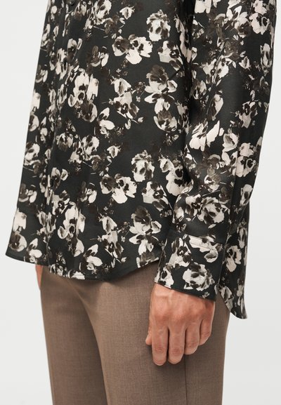 Μαύρο πουκάμισο μακρυμάνικο με λευκό και γκρι λουλουδάτο σχέδιο, φορεμένο με καφέ παντελόνια, δείχνοντας λεπτομέρειες στο πλάι και στο μανίκι.