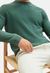 Grüner Pullover mit strukturierter Strickoptik und Rundhalsausschnitt; dazu cremefarbene Hose getragen. Sitzt auf einem Holzstuhl mit geflochtenem Sitz.