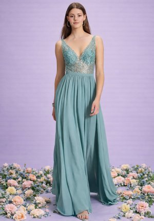 Junge Frau in einem ärmellosen Kleid in Türkis mit einem perlenbesetzten Oberteil steht barfuß zwischen verstreuten pastellfarbenen Blumen vor einem lavendelfarbenen Hintergrund.