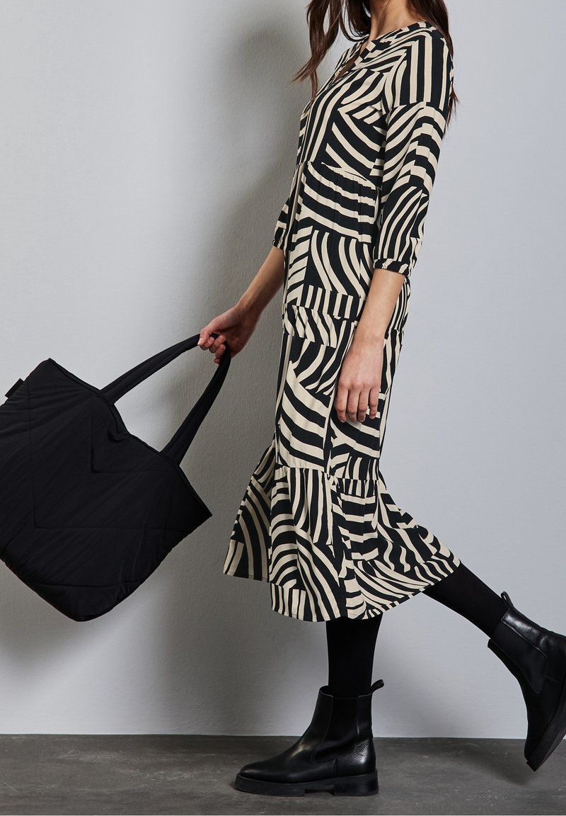 Femme en robe rayée noir et beige, collants noirs et bottes noires tenant un grand sac cabas noir, levant un pied en plein pas.