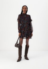 Vestido preto fluido com padrões paisley em vermelho e azul, mangas com babados e um decote alto. Combinado com botas de cowboy castanhas até ao joelho e uma bolsa.