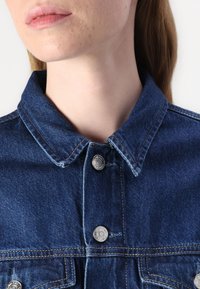 Giacca di jeans in tessuto blu, con collo a risvolto, bottoni in metallo argentato e dettagli di cucitura visibili.
