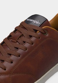 Καφέ δερμάτινο αθλητικό παπούτσι Wrangler με κορδόνια ίδιου χρώματος, μαύρη ετικέτα στη γλώσσα και ανοιχτό καφέ εσωτερική επένδυση σε λευκό φόντο.