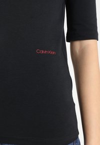 Černé tričko Calvin Klein s krátkými rukávy, hladká textura a červené logo na dolním levém rohu. Jednoduchý design, uvolněný střih.