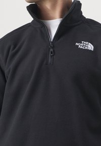 Černý fleecový svetr s polovičním zipem a bílým vyšívaným logem The North Face na levé hrudi. Měkká textura.