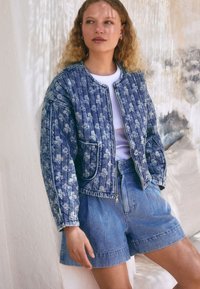 Denimjakke med blomstermønster, lynlåslukning foran og to lommer. Kombineret med højtaljede blå denimshorts, vist mod en neutral baggrund.