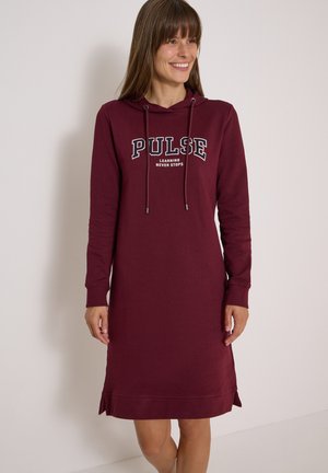 Frau trägt ein langärmliges kastanienbraunes Hoodie-Kleid mit dem Text "PULSE LEARNING NEVER STOPS", lächelnd und vor einem einfarbigen Hintergrund stehend.