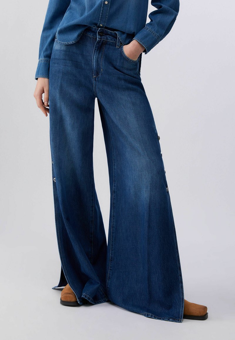 Person iført vidde jeans i mørkeblå med knapdetaljer på siderne, blå denimskjorte og brune sko, stående med den ene hånd i lommen.