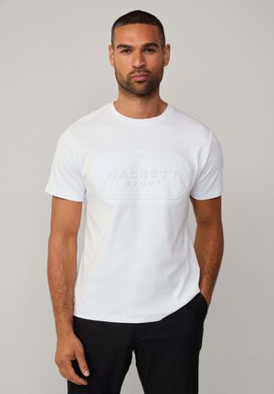 Camiseta de algodón blanca con cuello redondo, que presenta un logotipo texturizado en relieve que dice "HACKETT SPORT" en el frente. Llevada con pantalones negros.