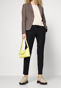 Blazer brun ajusté, haut beige, pantalon noir, sac à main jaune et chaussures beiges avec semelles épaisses noires. Tissus lisses et design structuré.