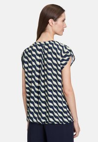 Granatowa bluzka krótki rękaw z falistym białym i jasnooliwkowym wzorem, o luźnym kroju i subtelnym detalem w postaci plisek na plecach.