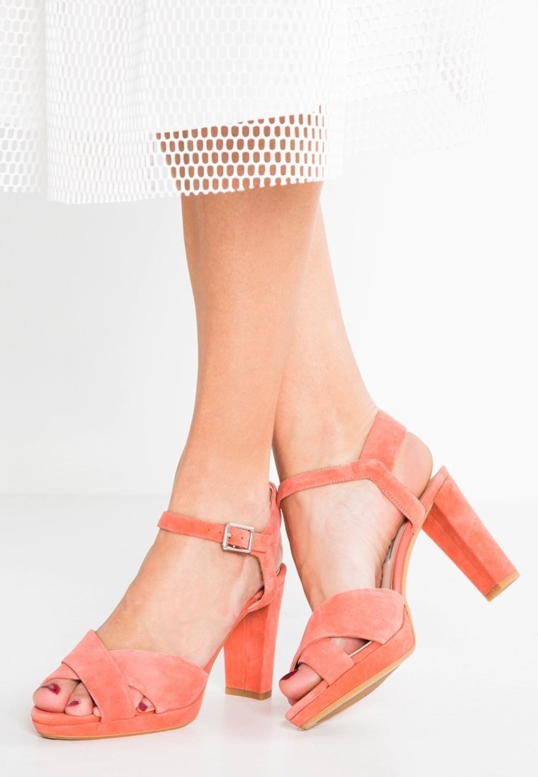 Des sandales à talons hauts en daim corail avec des sangles croisées à l'avant et une bride ajustable à la cheville. Talon carré et semelle en bois lisse.