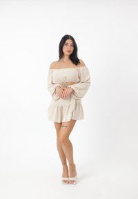 Beige Off-Shoulder Kleid mit langen, gepufften Ärmeln, tailliertem Bund und gestuften Saum; getragen mit weißen Riemchenschuhen.