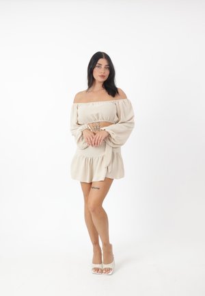 Beige Off-Shoulder Kleid mit langen, gepufften Ärmeln, tailliertem Bund und gestuften Saum; getragen mit weißen Riemchenschuhen.