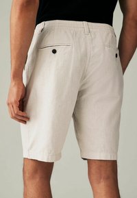 Homme portant un short beige clair avec une taille élastique et des poches arrière boutonnées, se tenant devant un fond neutre.