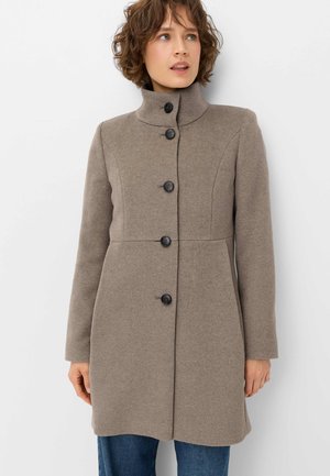 Woman wearing a taupe buttoned wool coat with a high collar, standing against a plain white background, looking slightly to the side.