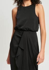 Vestido negro sin mangas con una tela suave y fluida. Presenta una cintura fruncida y un detalle drapeado en la parte frontal para añadir dimensión.