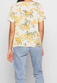Kurzärmliges, hellbeiges Oberteil mit gelbem Blumenmuster und grünen Akzenten, mit rundem Ausschnitt und lässiger Passform. Getragen mit hellblauen Jeans.