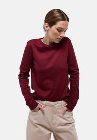 Chemise bordeaux à manches longues avec un col rond, confectionnée dans un tissu lisse. Associée à un pantalon beige clair doté de poches latérales.