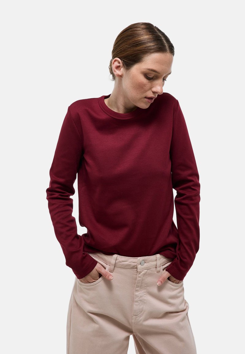 Chemise bordeaux à manches longues avec un col rond, confectionnée dans un tissu lisse. Associée à un pantalon beige clair doté de poches latérales.