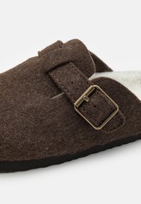 Papuci maro din fetru, cu o curea cu cataramă, căptușiți cu un material moale și cu o talpă neagră. Textura pare pufoasă, având un design cu tăietură dreaptă.