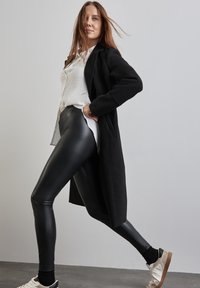 Cappotto lungo nero su una camicia bianca, abbinato a leggings in ecopelle neri e sneakers chiare. Lo sfondo è grigio chiaro e il pavimento è grigio scuro.