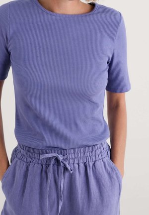 Person trägt ein violettes, geripptes Kurzarm-Top und passende violette Leinenhose mit Kordelzug, Hände in den Taschen.