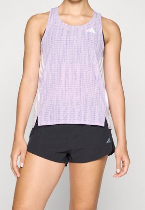 Frau trägt ein ärmelloses lavendelfarbenes Sporttop mit weißem Logo und schwarze Laufshorts vor einfarbigem Hintergrund.