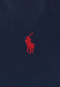 Tmavě modrá texturovaná látka s výrazným červeným vyšívaným logem hráče póla, umístěným uprostřed materiálu pro jasnou viditelnost.