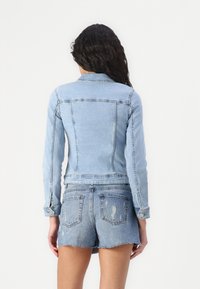 Kvinna med långt mörkt hår som bär en ljusblå denimjacka och slitna denimshorts, stående vänd bort från kameran mot en vit bakgrund.
