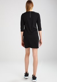 Svart klänning med trekvartslånga ärmar, dragkedjedetalj på ryggen och en figurnära silhuett, kombinerad med svarta sportskor. Slät textur.