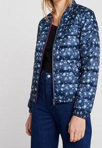 Navy pufferjack met een bloemenpatroon, ritssluiting aan de voorkant, ribgebreide manchetten en zoom. Gestyled met een zwarte top en high-waisted denim jeans.