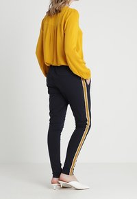 Blouse jaune à manches longues avec un pli dans le dos, associée à un pantalon slim noir avec des rayures blanches et dorées sur les côtés, et des mules blanches à talons.