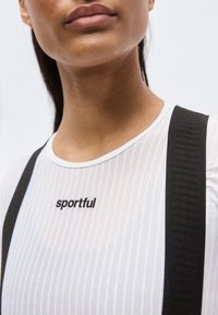 Κοντινό πλάνο ενός ατόμου που φοράει ένα λευκό ριγωτό τοπ με το κείμενο "sportful" και μαύρες τιράντες, σε απλό ανοιχτό φόντο.