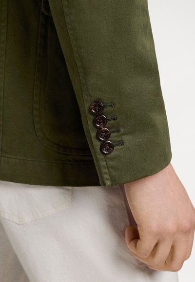 Κοντινό πλάνο του χεριού ενός ατόμου δίπλα στο μανίκι μιας ελιάς πράσινης ζακέτας με τέσσερα σκούρα καφέ κουμπιά, σε συνδυασμό με παντελόνι ανοιχτού χρώματος.