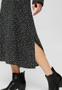 Černé midi šaty s bílým puntíkovým vzorem a bočním rozparkem. Nošeny s černými kotníkovými botami. Hladká látka a rozšířený střih.